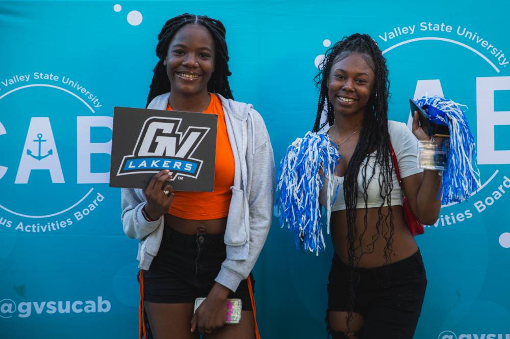 two people posing in front of CAB backdrop at Laker Kickoff photo booth and holding GV sign and blue and white pom poms